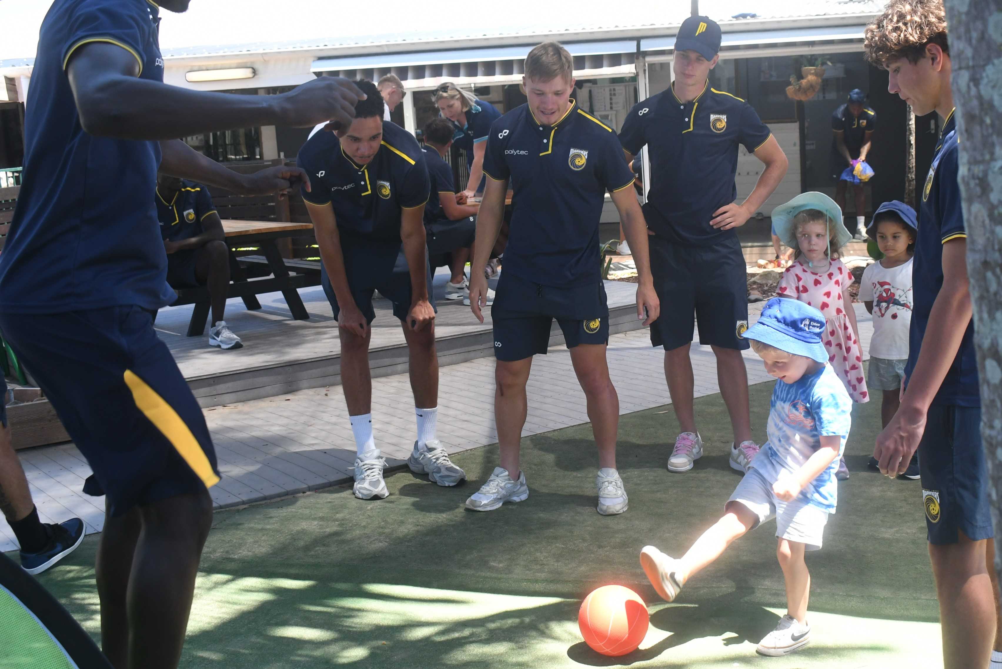 Players from the Central Coast Mariners watch as a child kicks a football.